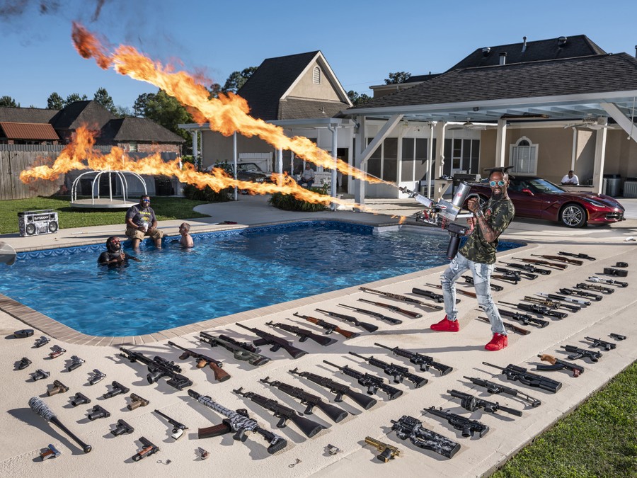 A man stands near a backyard swimming pool, surrounded by dozens of weapons laid out on the concrete, firing two flamethrowers.