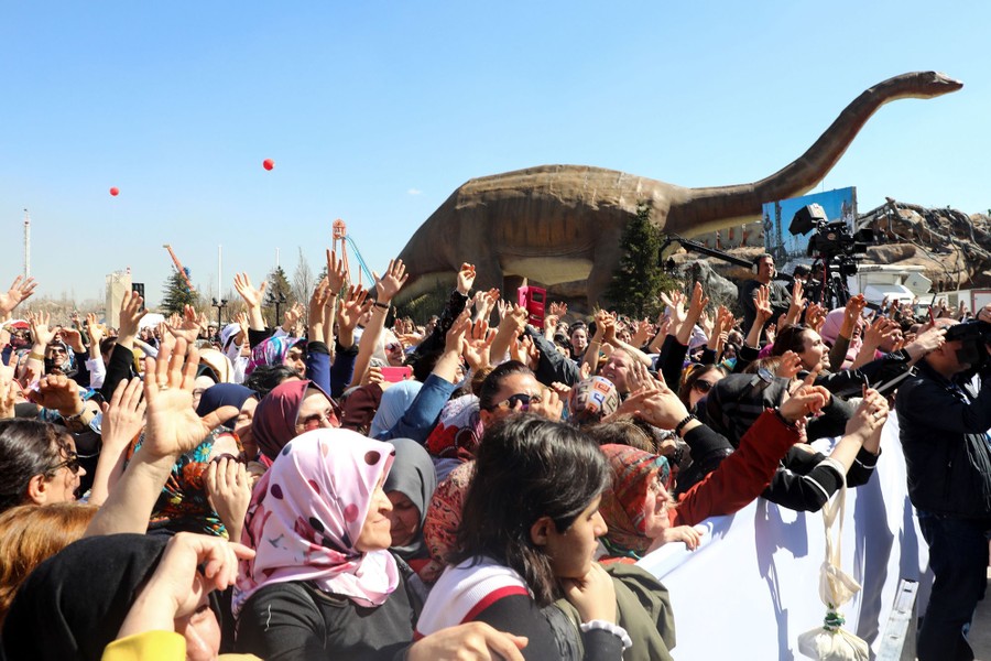 A crowd of people cheer in front of a large statue of a dinosaur.