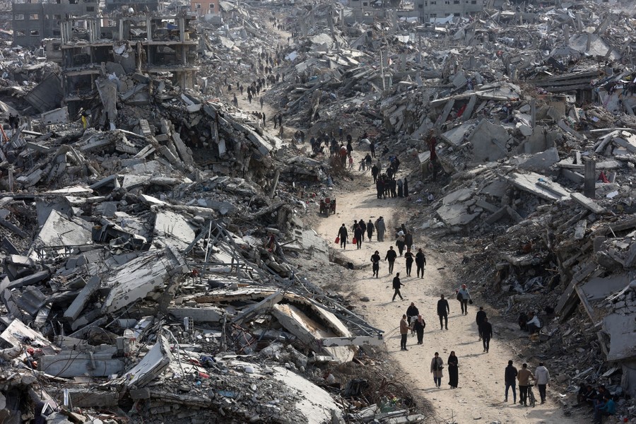 People walk in small groups down a road surrounded by many immense piles of rubble and debris.
