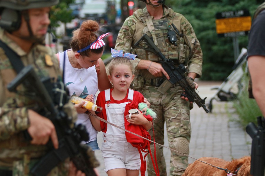 Several armed officers walk beside civilians, including a young girl, who appears distressed.