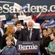 Bernie Sanders, standing at a lectern, surrounded by his family, laughs as he looks onto a crowd.