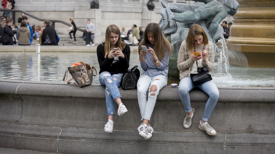 Three teenage girls sitting side by side on a fountain in Trafalgar Square are lost in their phones