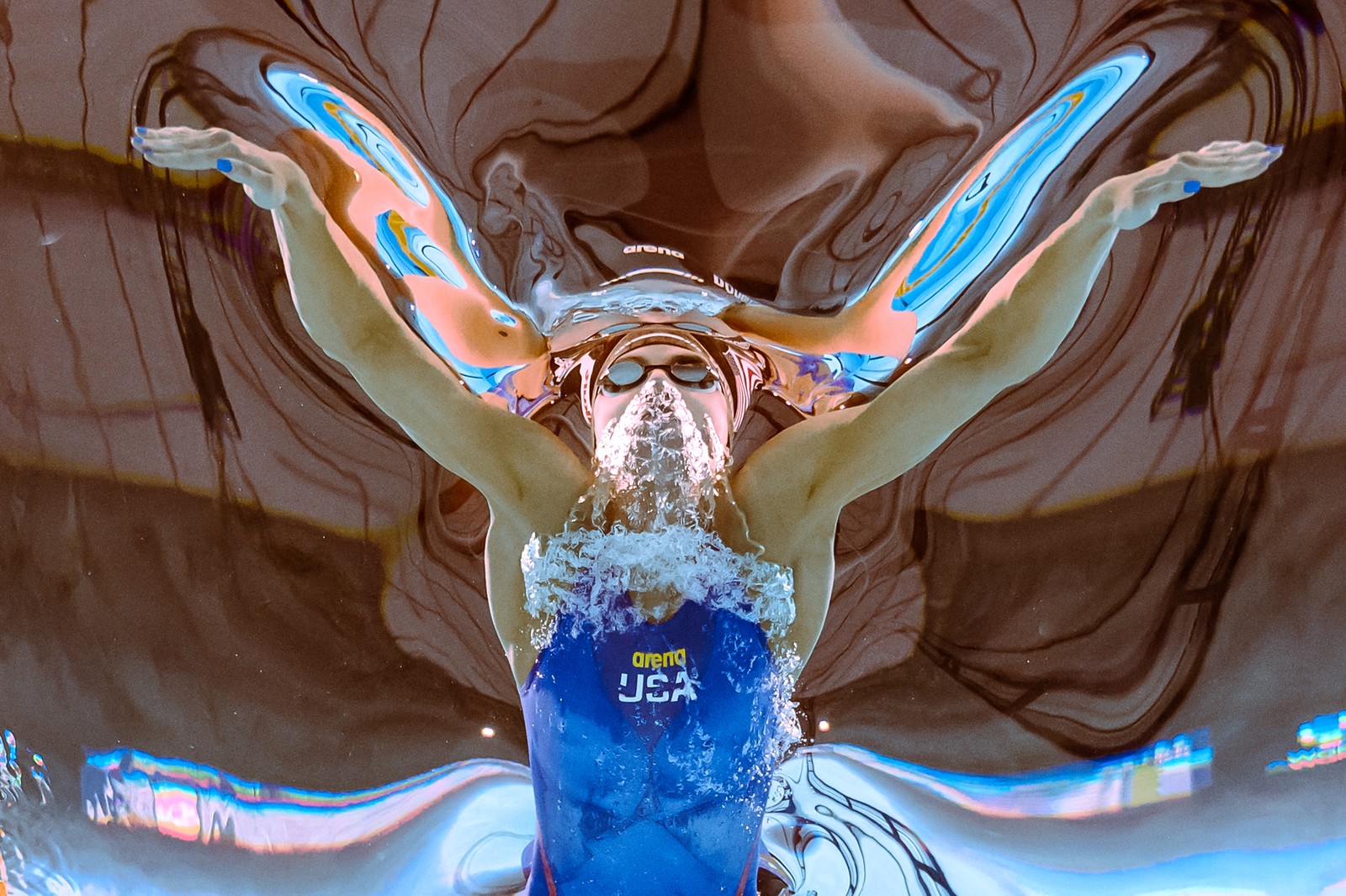 An underwater view of a swimmer passing by overhead, creating waves and distorting the view of the ceiling beyond.