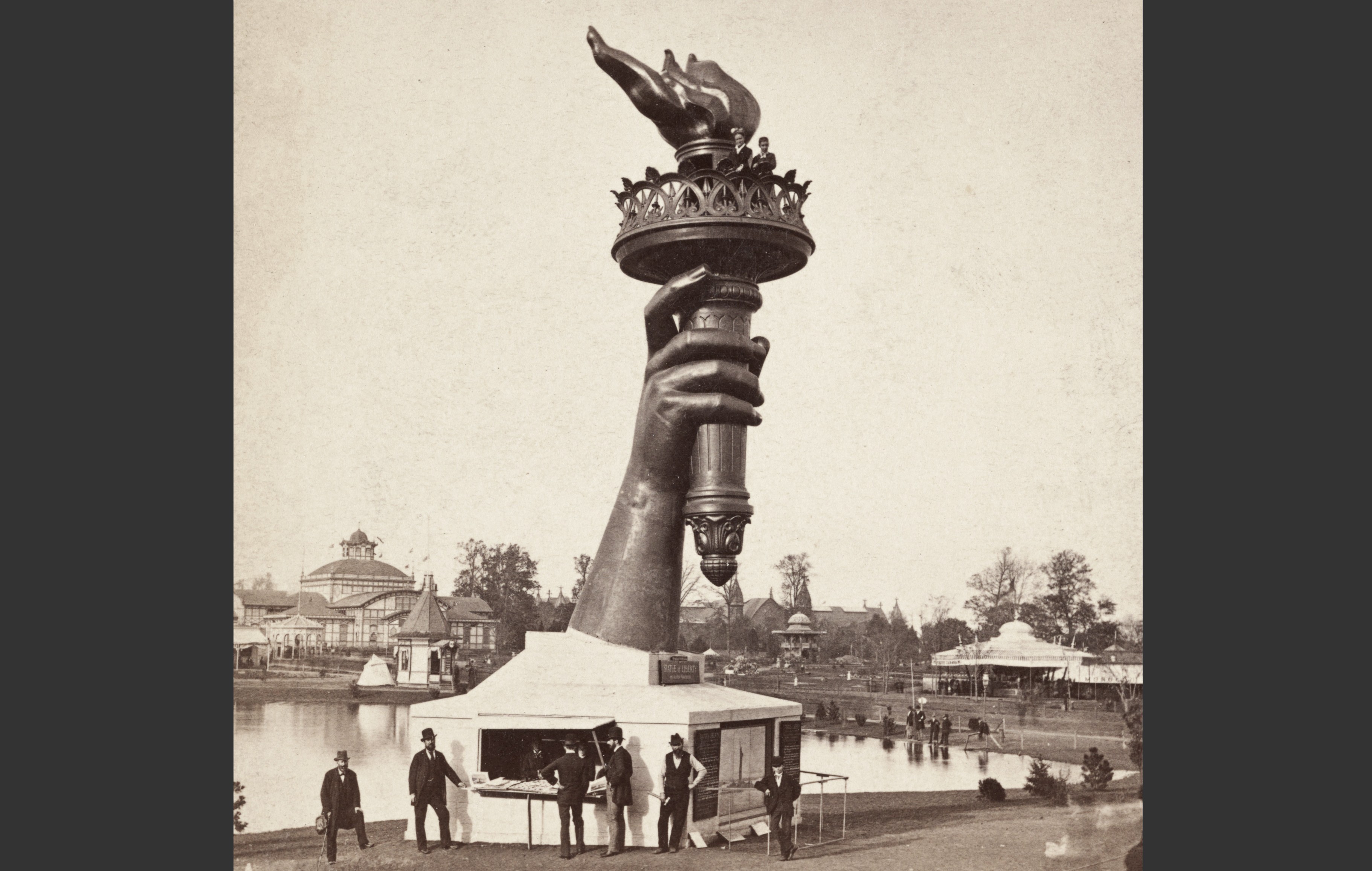 People gather at an information booth at the base of a very tall statue of an arm holding a torch, in a photo from 1876.