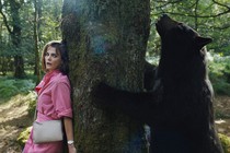A film still of Cocaine Bear standing on one side of a tree, and a woman hiding on the other