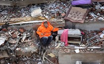 A man sits in the rubble of a collapsed building, his left arm reaching out and holding the hand of his daughter, who lies dead, trapped beneath the debris.