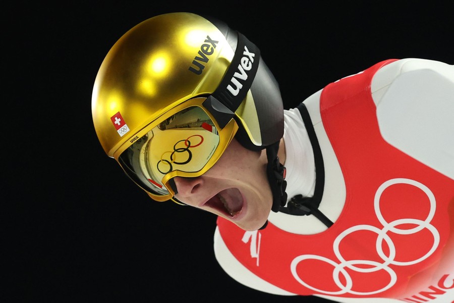 A ski jumper is seen in action, mouth wide open; a steep slope ahead is seen reflected in his visor.