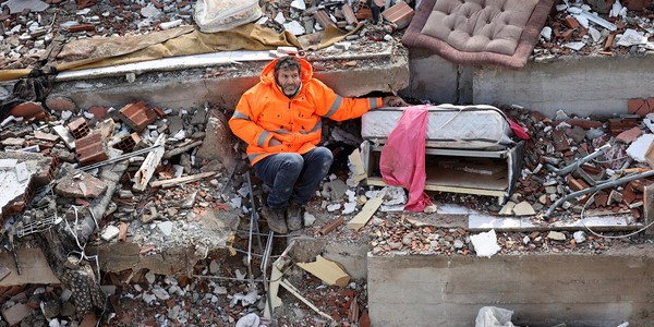 A man sits in the rubble of a collapsed building, his left arm reaching out and holding the hand of his daughter, who lies dead, trapped beneath the debris.