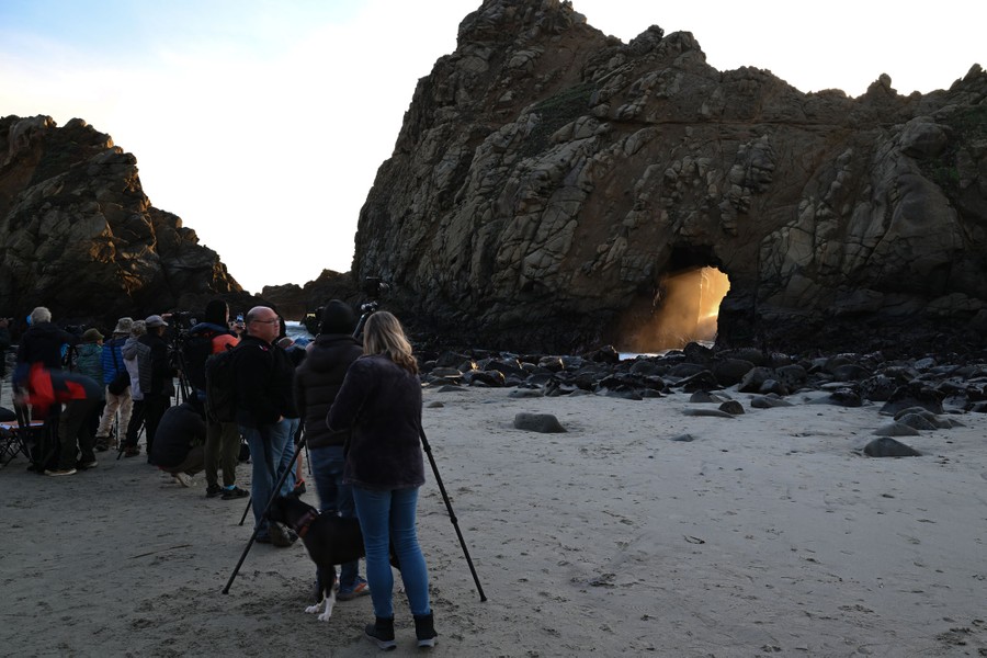 A group of people stand on a beach, taking photographs of sunlight streaming through an arch in a stone outcrop.