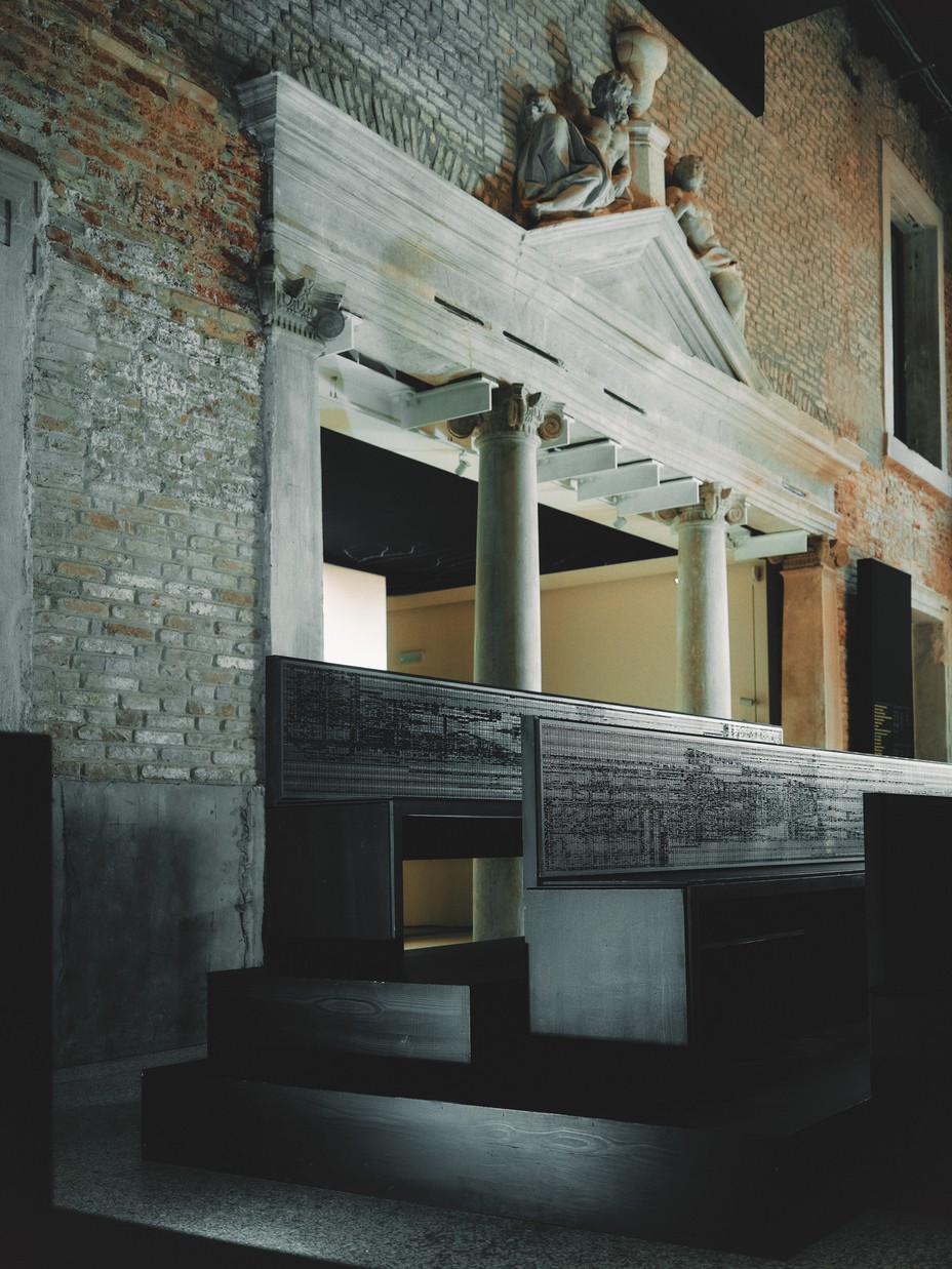 Image 4: photo of installation with dark stair-stepped wooden benches in large brick hall with marble pediment and sculptures over wide doorway with marble columns