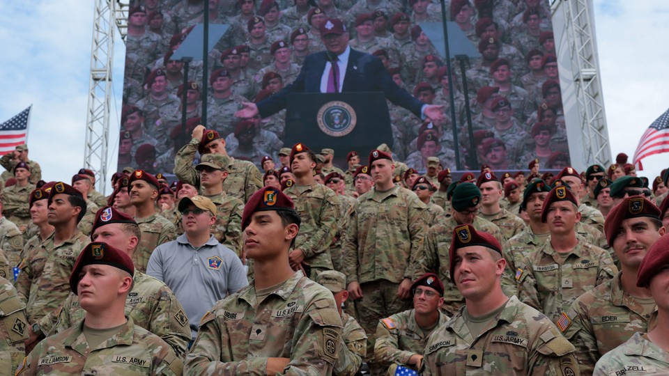 A group of soldiers stands in front of a Jumbotron showing Donald Trump at a podium above them