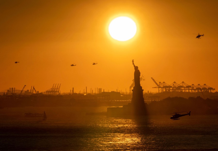 Helicopters fly in the distance as the sun sets behind the Statue of Liberty.