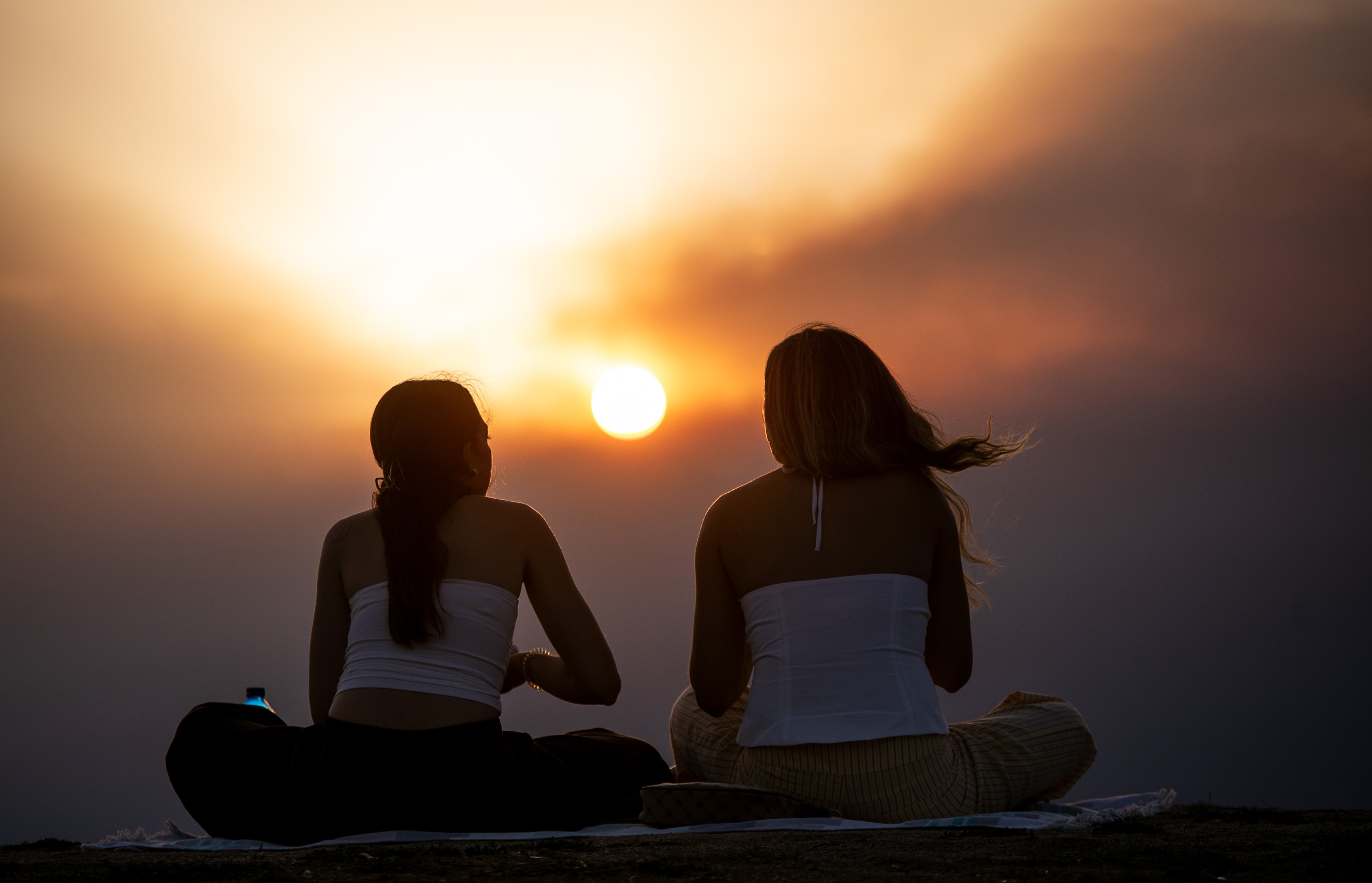 Two people sit outside, watching a sunset, under smoky skies.