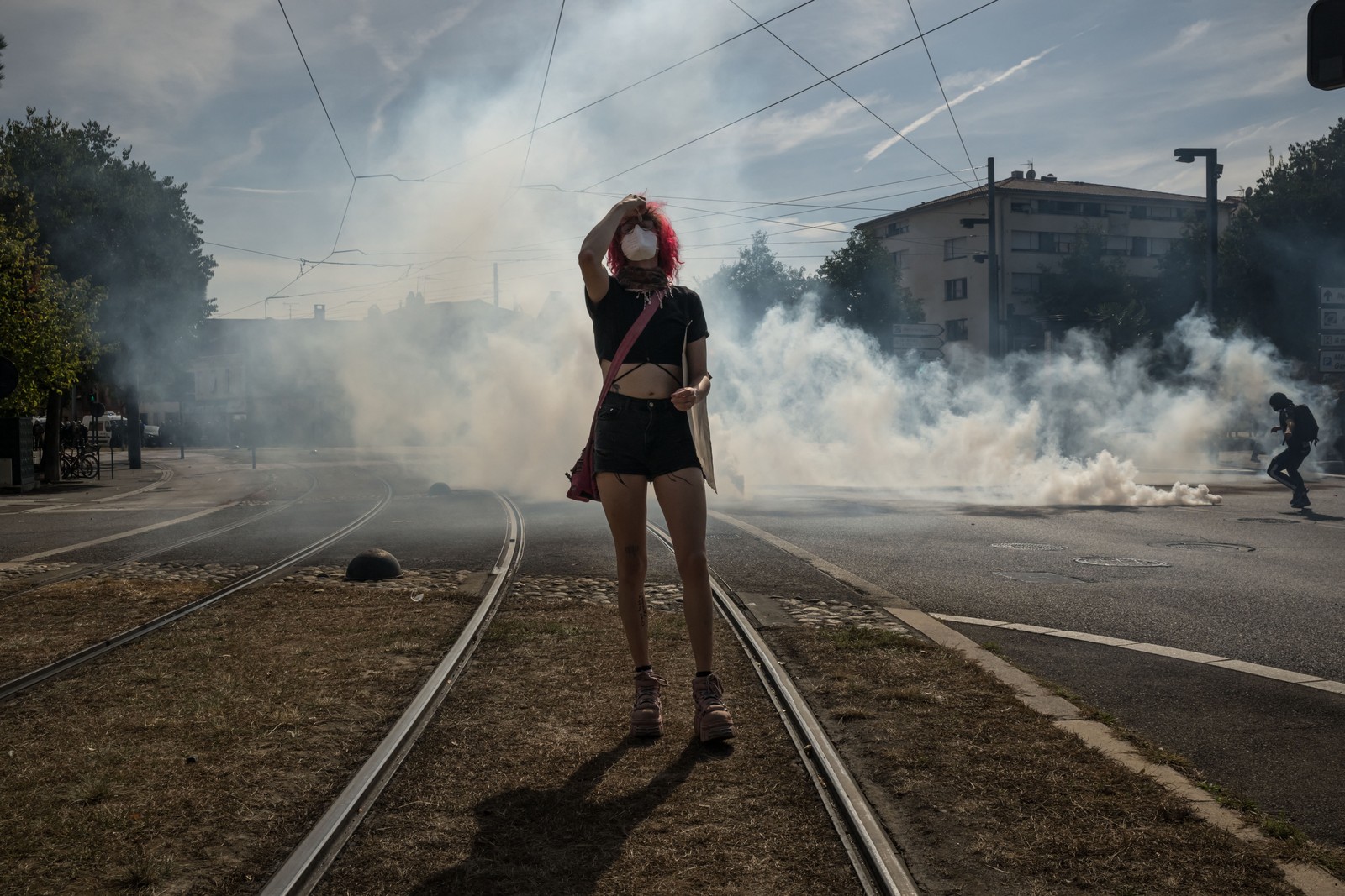 A student protester wearing a face mask walks, holding their head; cloud of tear gas is in the background.