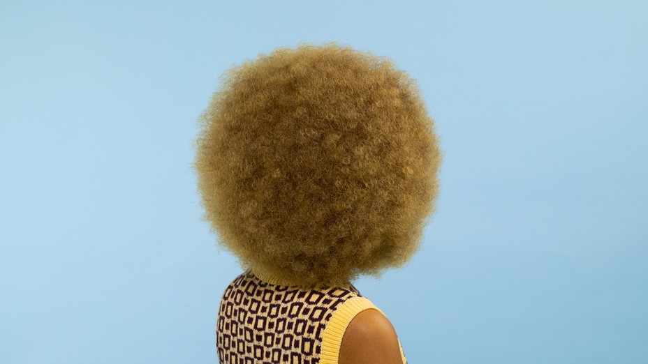 woman with blond afro from behind wearing yellow on blue background