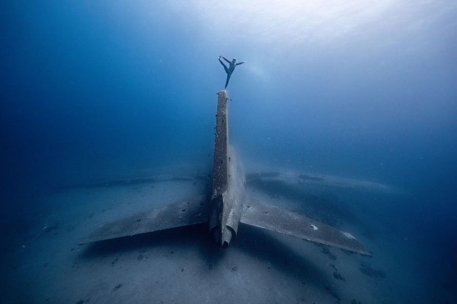 A diver poses while "standing" underwater, atop the tail fin of a sunken aircraft.