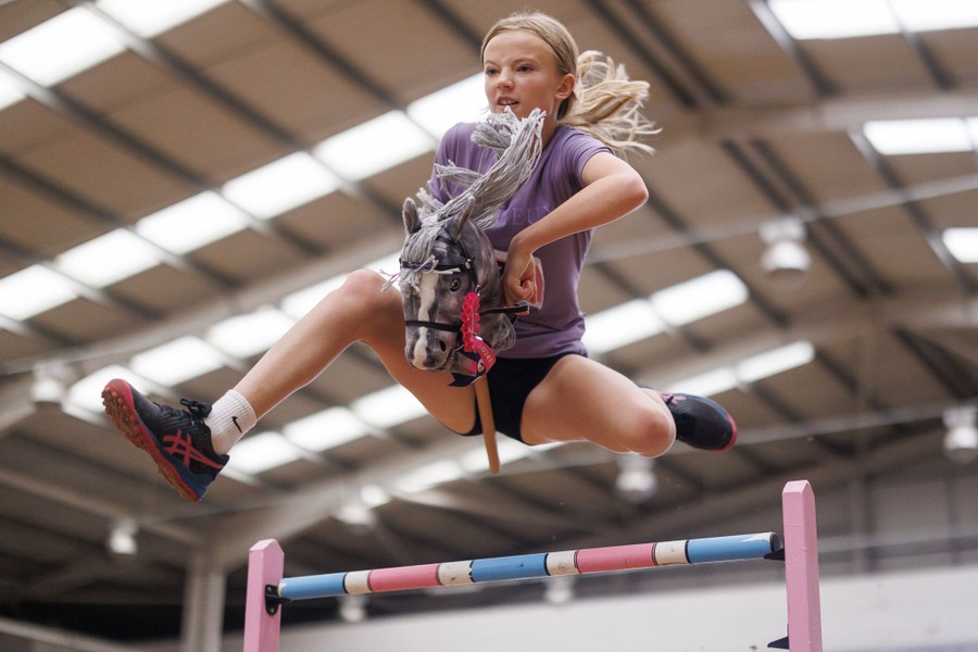 A young person leaps over an obstacle while holding a hobbyhorse.