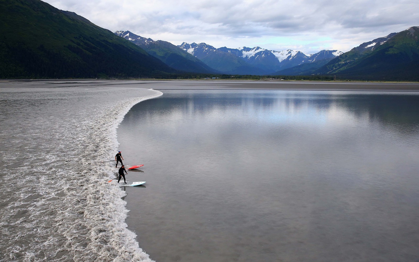 A pair of surfers catch a bore tide—a wide single wave flowing through a relatively narrow and shallow channel, seen among nearby mountains.
