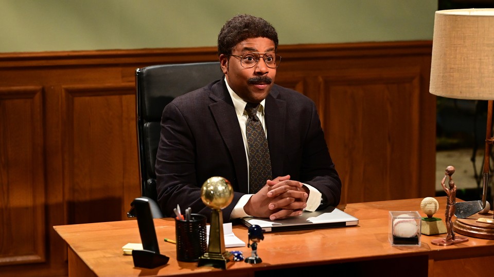 Kenan Thompson, wearing oversize glasses and a mustache, sits behind a desk with hands folded.