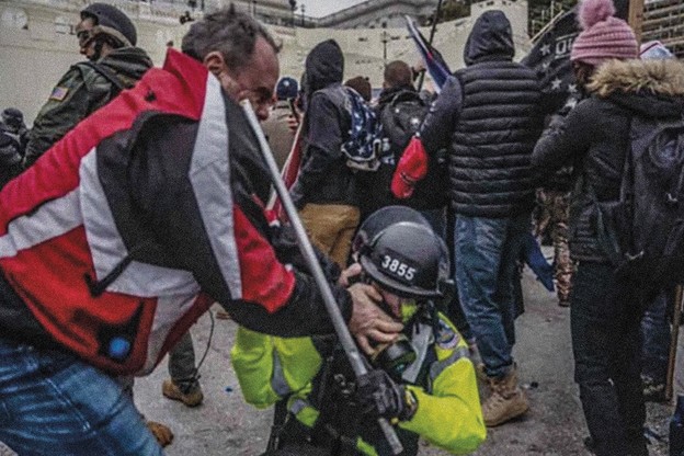 photo of man in red, gray, black jacket and jeans standing over and pushing his thumbs into the eyes of a kneeling police officer in helmet and gear