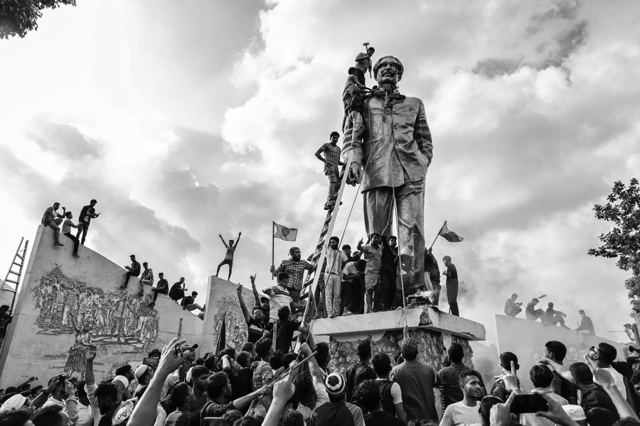 A group of protesters surround and climb on a tall statue of the former president, with one holding a sledgehammer atop a ladder.