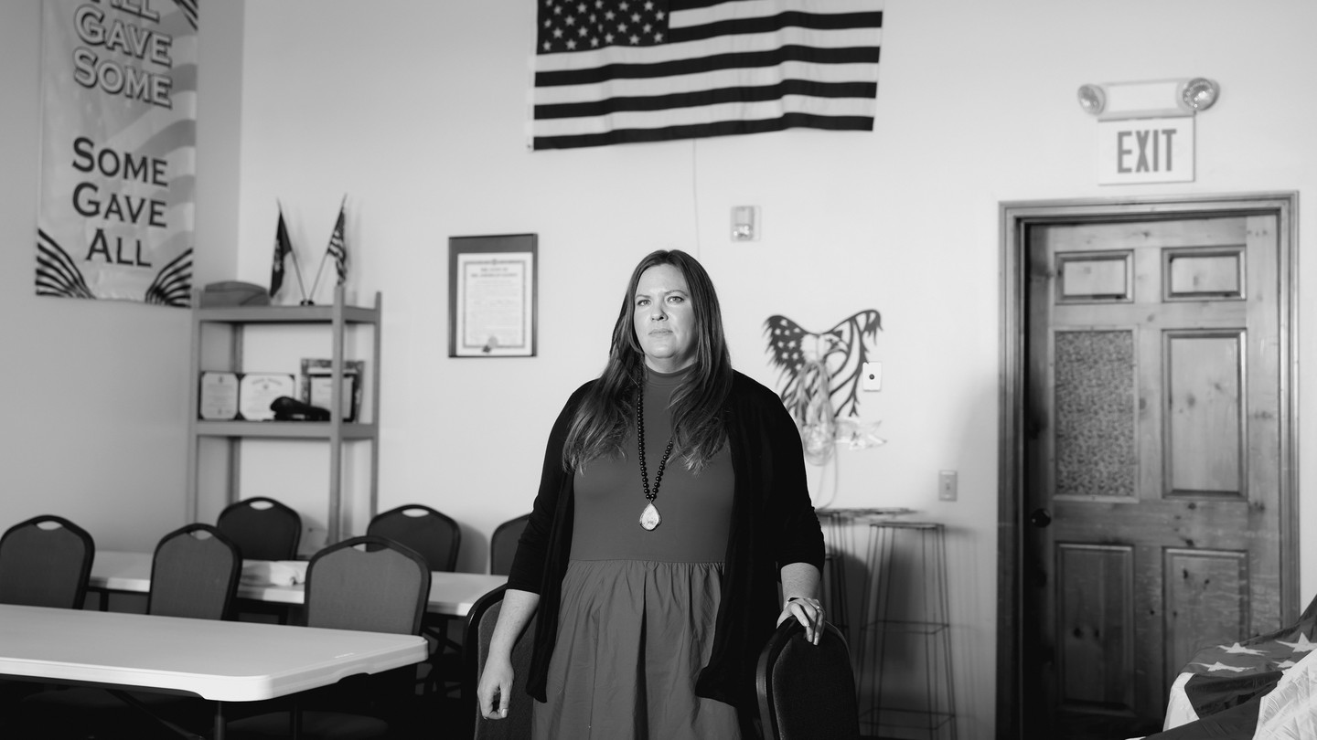 A black-and-white photo of a white woman standing in a room with the American flag behind her