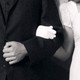 A black-and-white photo of man in a tux and a woman in a white dress, with arms locked