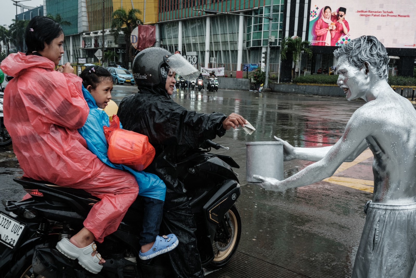 A performer covered in silver paint receives a tip from a person on a scooter in a street.