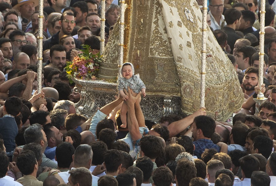 People in a large crowd hold a young child up near a religious statue being carried.