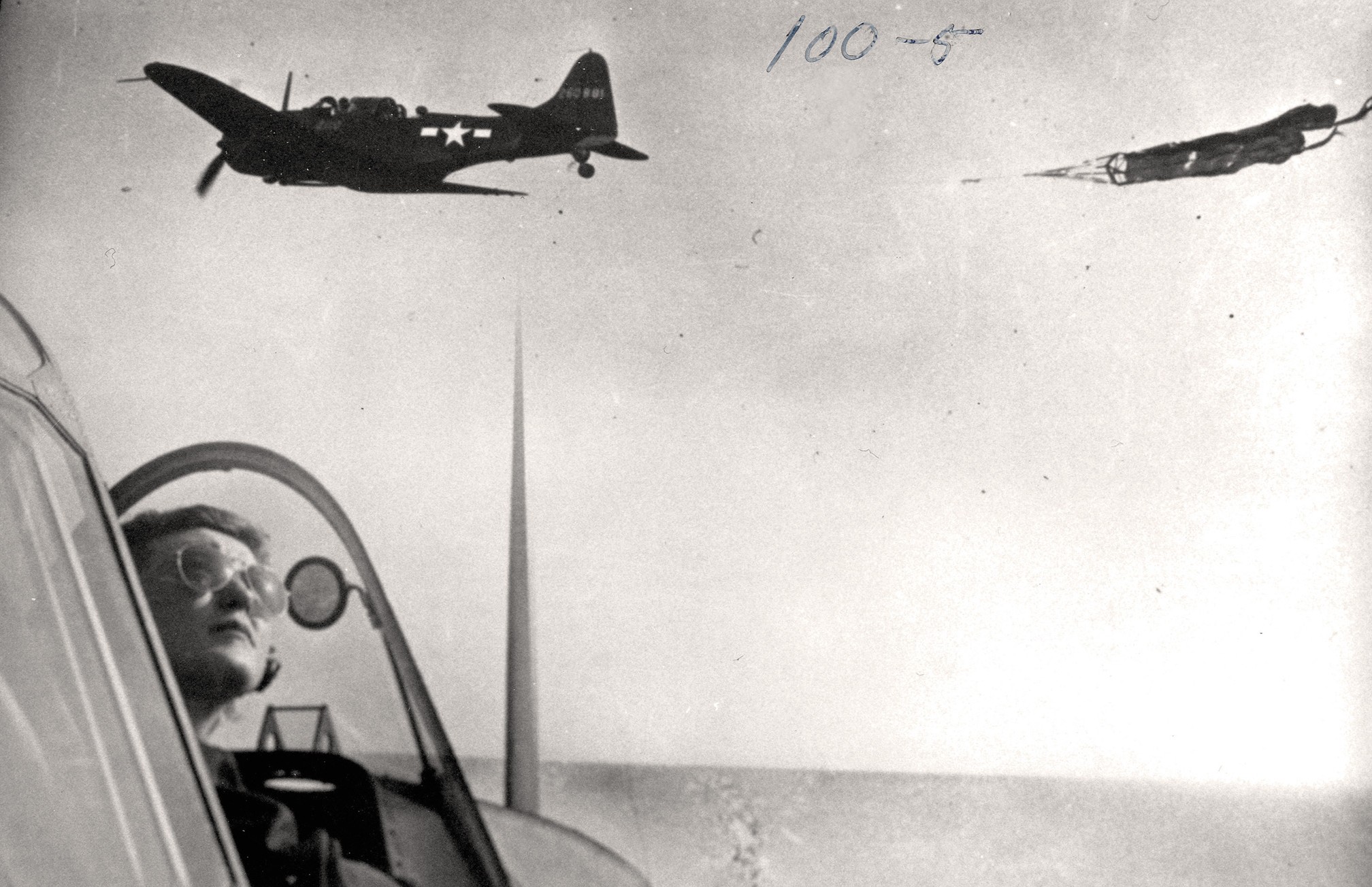 black-and-white archival photo of woman wearing aviators in open cockpit looking up as another plane towing a target passes perpendicularly overhead