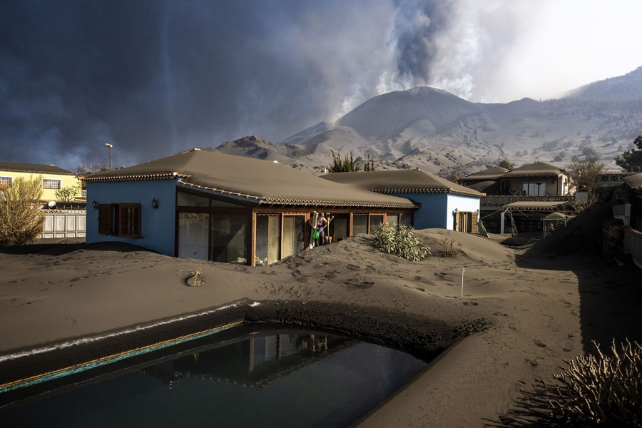 A person walks out of their ash-covered house as a volcano erupts in the background.