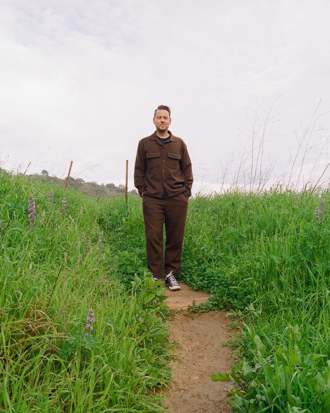 Color photo of man in brown shirt and pants standing with hands in pockets on trail surrounded by green grass and wildflowers.