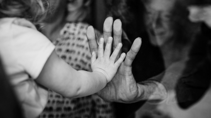 A little girl presses her hand to an old woman's through a glass window
