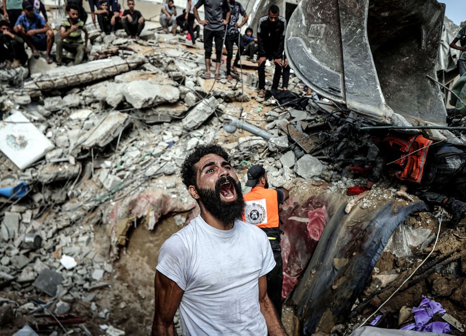A man reacts, standing beside a huge pile of rubble following an air strike. Workers and onlookers stand nearby and atop the rubble.