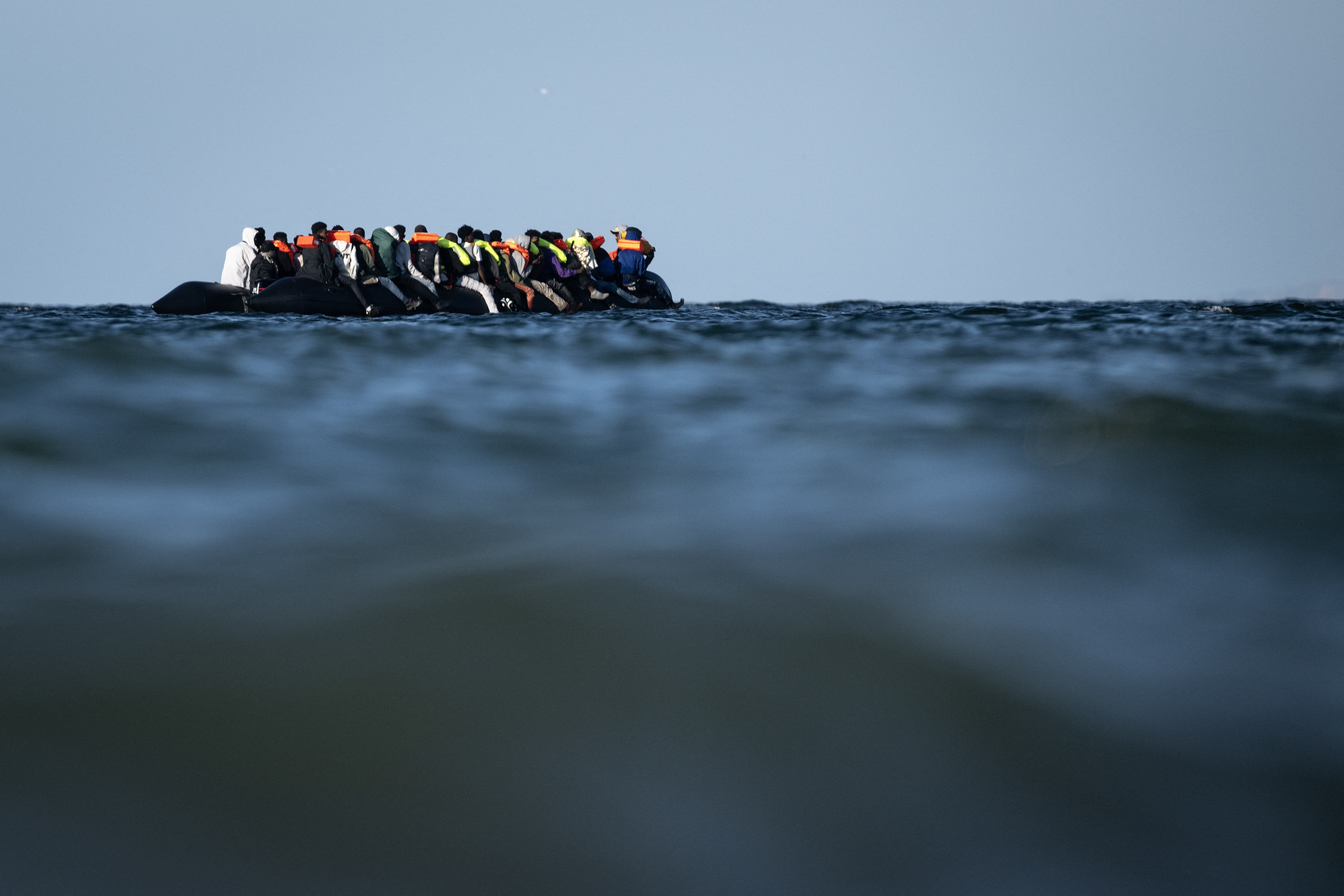 A low-angle view of many migrants wearing life vests sitting on an inflatable raft in the ocean