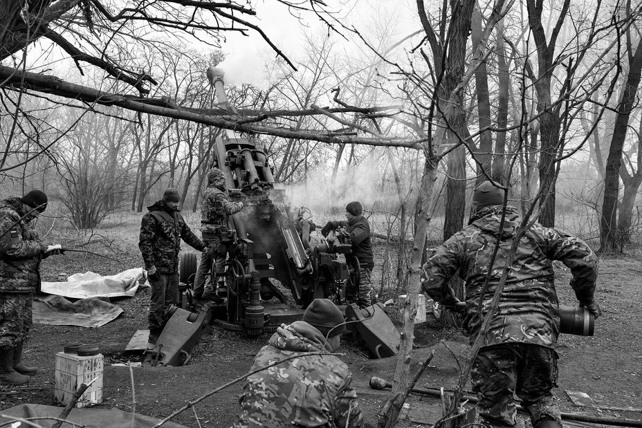 black-and-white photo of group of soldiers in forest firing howitzer with a soldier carrying the next shell to load
