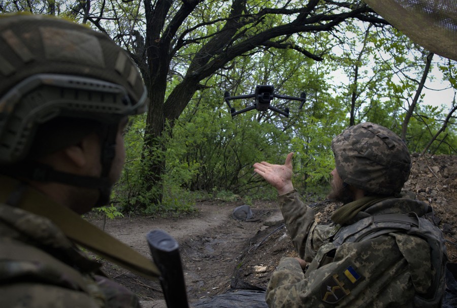 Two soldiers fly a small drone among trees.