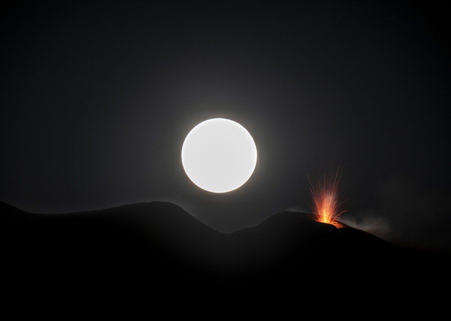 A full moon rises behind a small volcanic eruption atop a large volcano.
