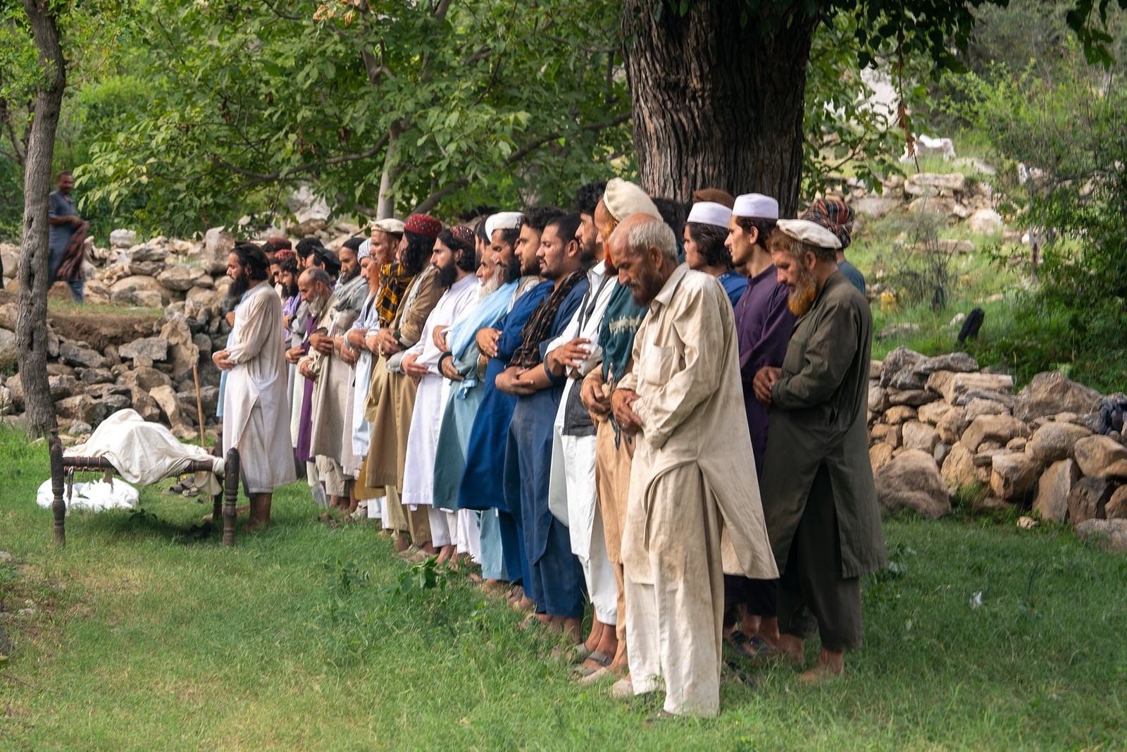 Men stand side-by-side outside, praying during a funeral ceremony, standing in front of several bodies wrapped in sheets.