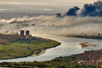 An elevated view of a river running through a city with black and white smoke rising from numerous fires.