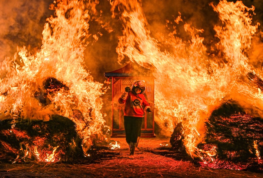 Several people carry an ornamental wooden box past large bonfires.