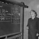 A black and white photograph of Albert Einstein in front of a blackboard