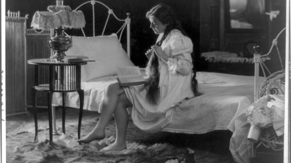 A young woman reads at the corner of her bed as she braids her hair.