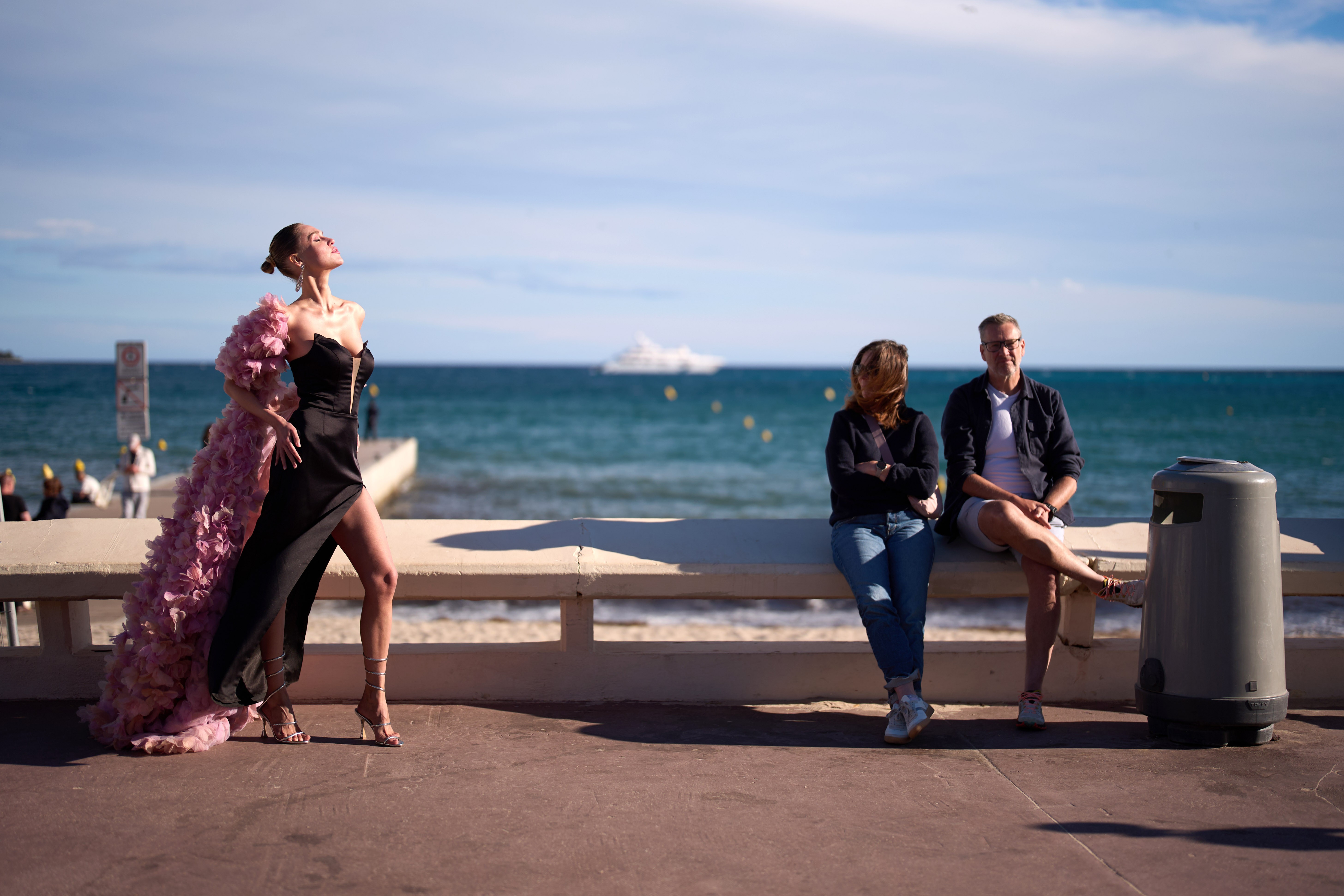 A person in a formal dress poses for a photo, beside visitors casually sitting on a seawall nearby.