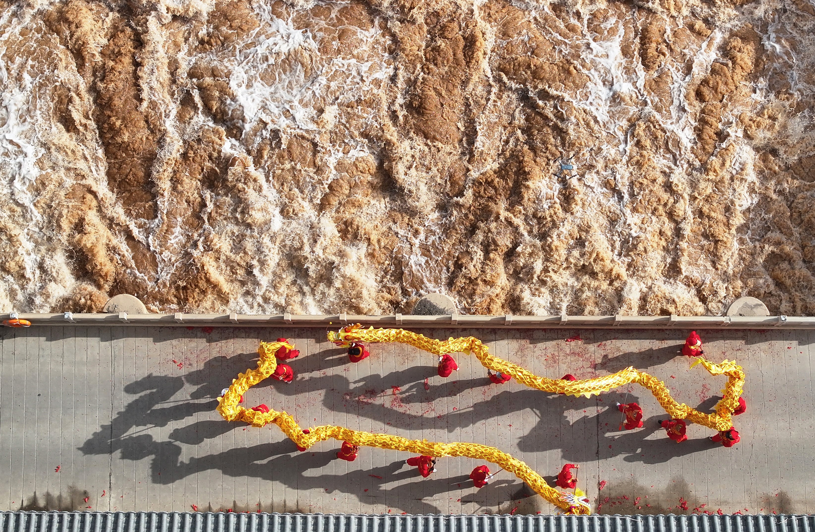 A drone photo shows roiling muddy water flowing from an opened sluice gate, alongside a platform where people perform a dragon dance.