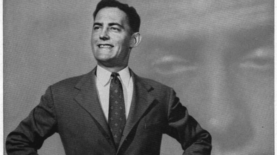 A black-and-white photo of a smiling man in a suit and tie with his hands at his hips