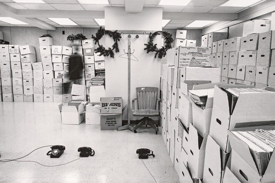 storage room with dropped ceiling, fluorescent lights, holiday decorations, 3 rotary-dial phones in the middle of a bare floor, and dozens of white file boxes stacked along walls