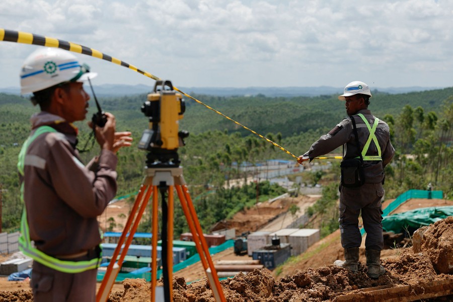 Two construction workers stand on a hillside beside surveying equipment.
