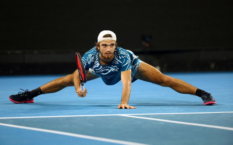 A tennis player leans low to the ground, legs spread wide, during a match.