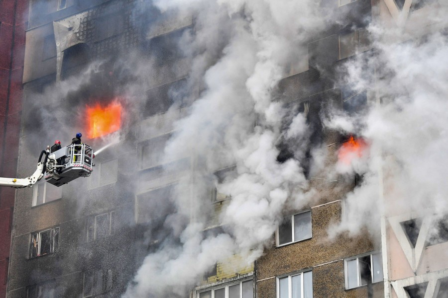 Firefighters stand in a basket at the end of a long lift, spraying water onto a tall residential building that is on fire.